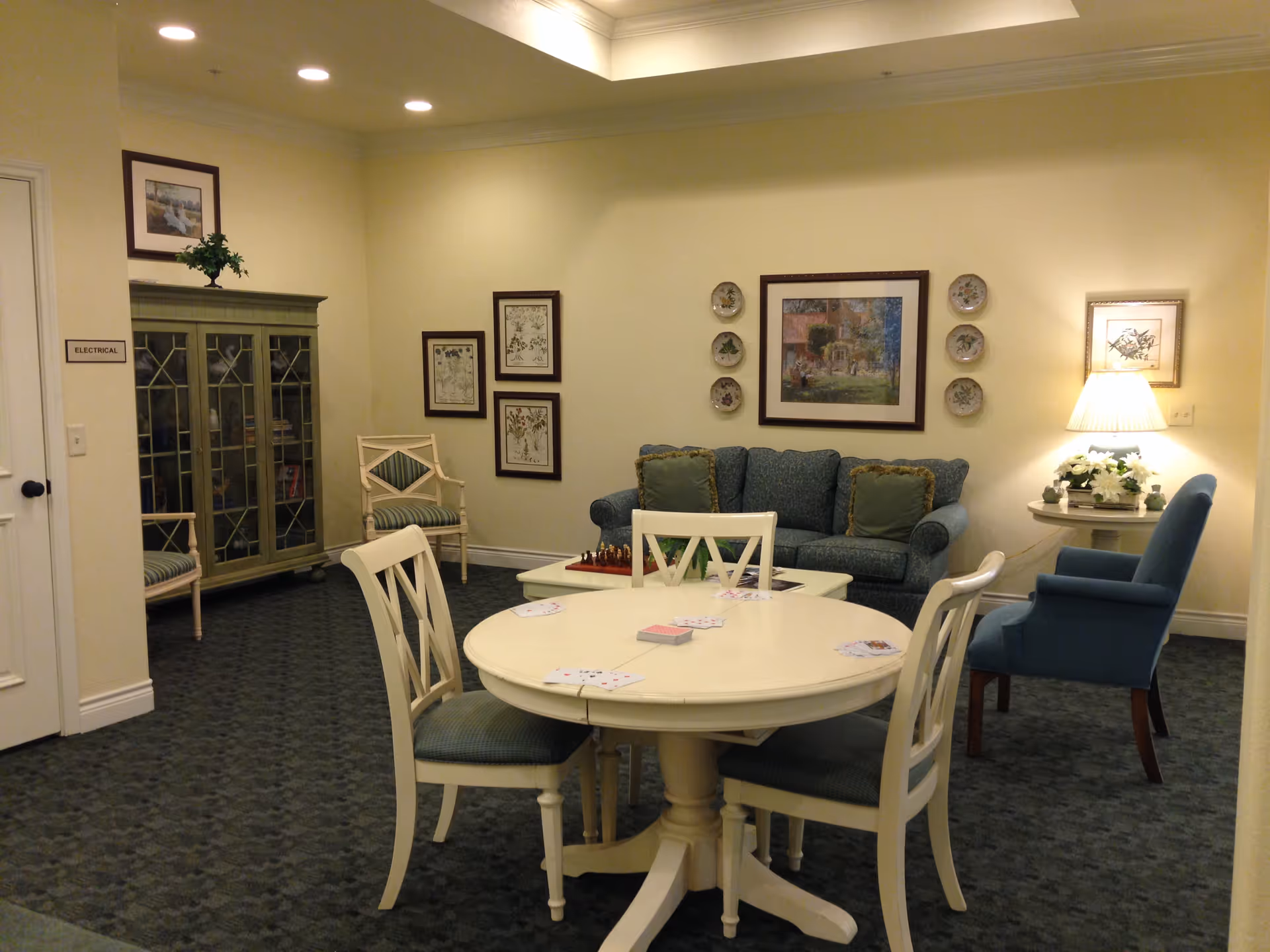 A cozy common area with a round table and four chairs in the foreground, playing cards spread on the table. In the background, there is a blue couch with green pillows, a blue armchair, a side table with a lamp and flowers, and framed artwork on the walls. A glass-front cabinet and additional chairs are also visible.