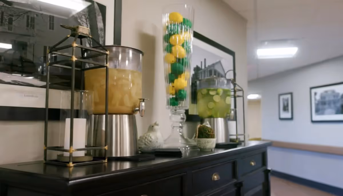 A dark wooden sideboard with two beverage dispensers filled with yellow and green drinks, a tall glass container filled with decorative yellow and green balls, a white ceramic bird figurine, and a candle lantern. The background shows a hallway with framed black and white photographs on the walls.