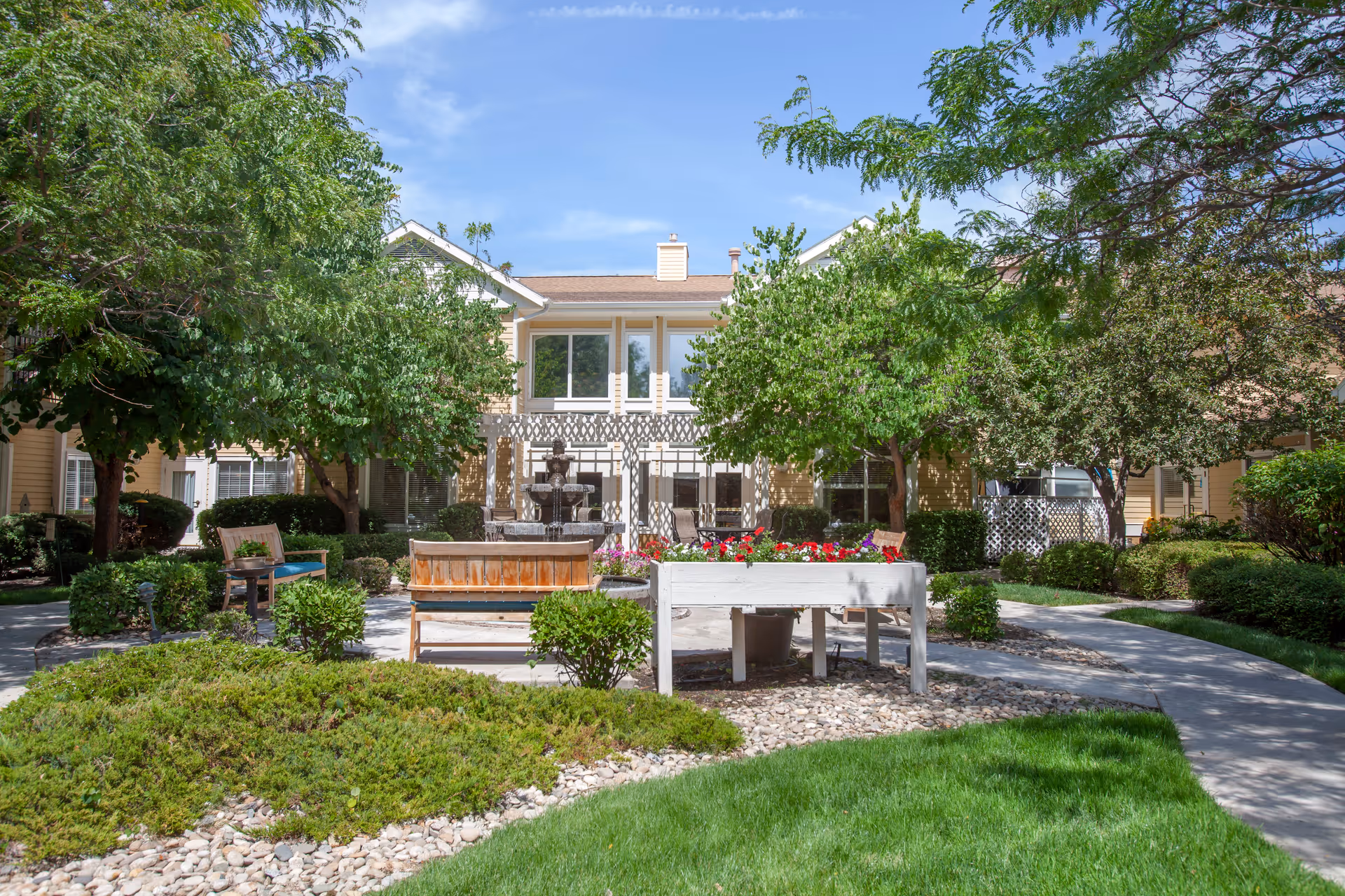 Outdoor courtyard with benches, a planter of flowers, a central fountain, trees, and the facility building behind.
