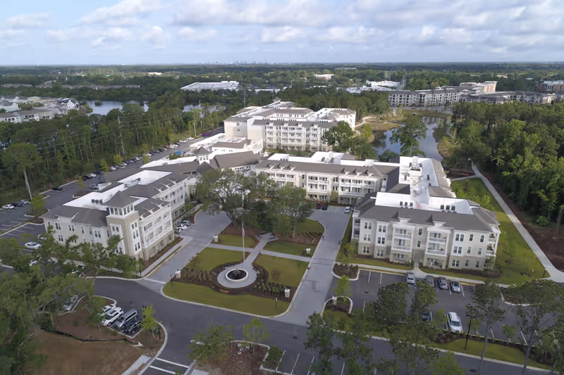 Aerial view of South Bay At Mount Pleasant senior living facility showing multiple connected buildings surrounded by trees, parking lots, and landscaped areas with a circular driveway and flagpole in the center.