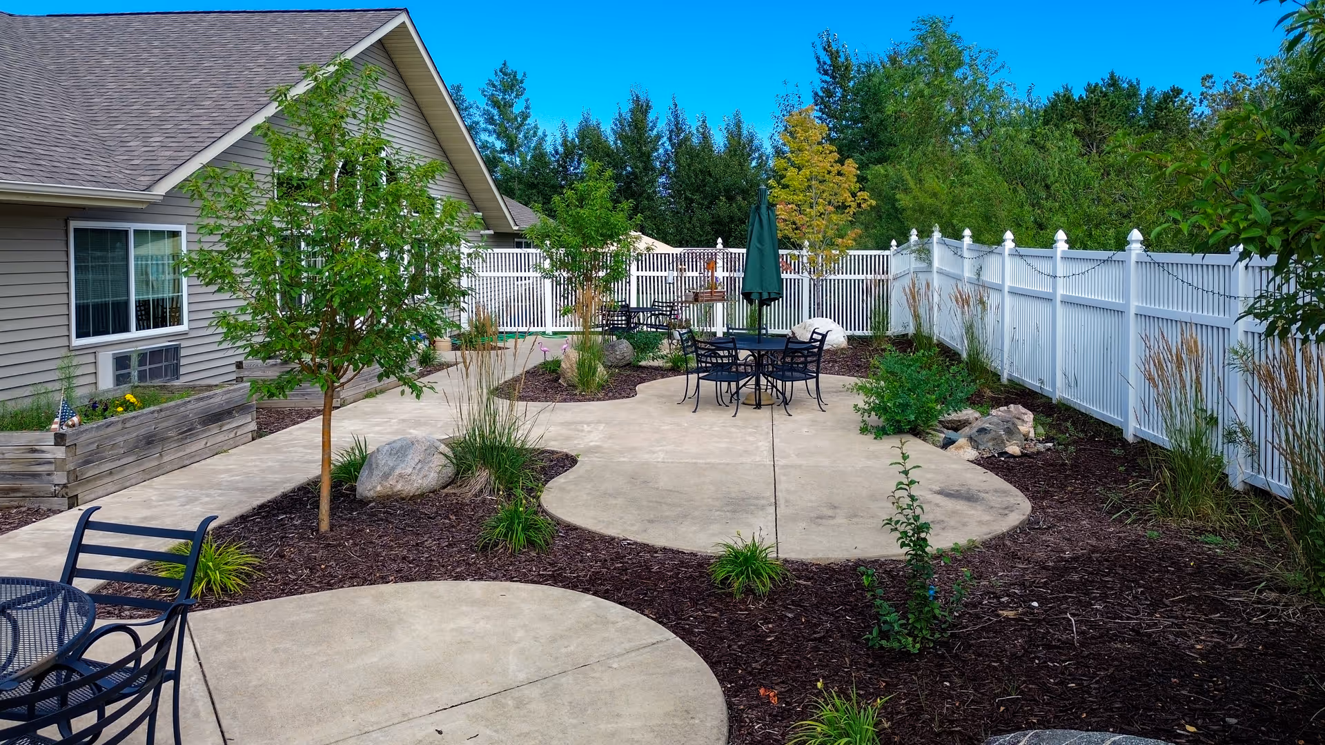 Outdoor patio area with concrete walkways, metal tables and chairs, small trees, plants, and a white fence surrounding the space. The side of a beige building with windows is visible on the left.