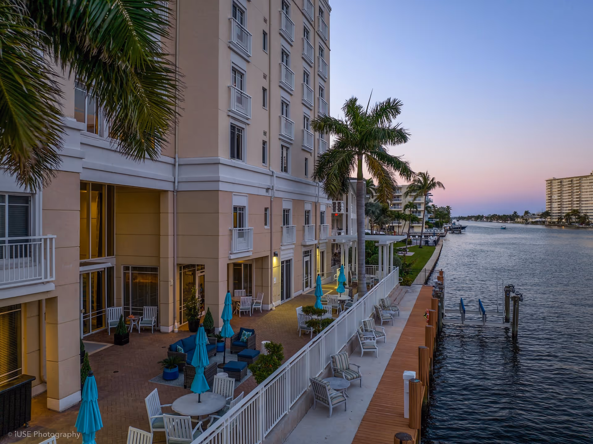 Outdoor patio area of a senior living facility with seating arrangements including chairs, tables with umbrellas, and lounge chairs along a waterfront. Palm trees and a multi-story building are visible, with a calm waterway extending into the distance under a clear sky at dusk.