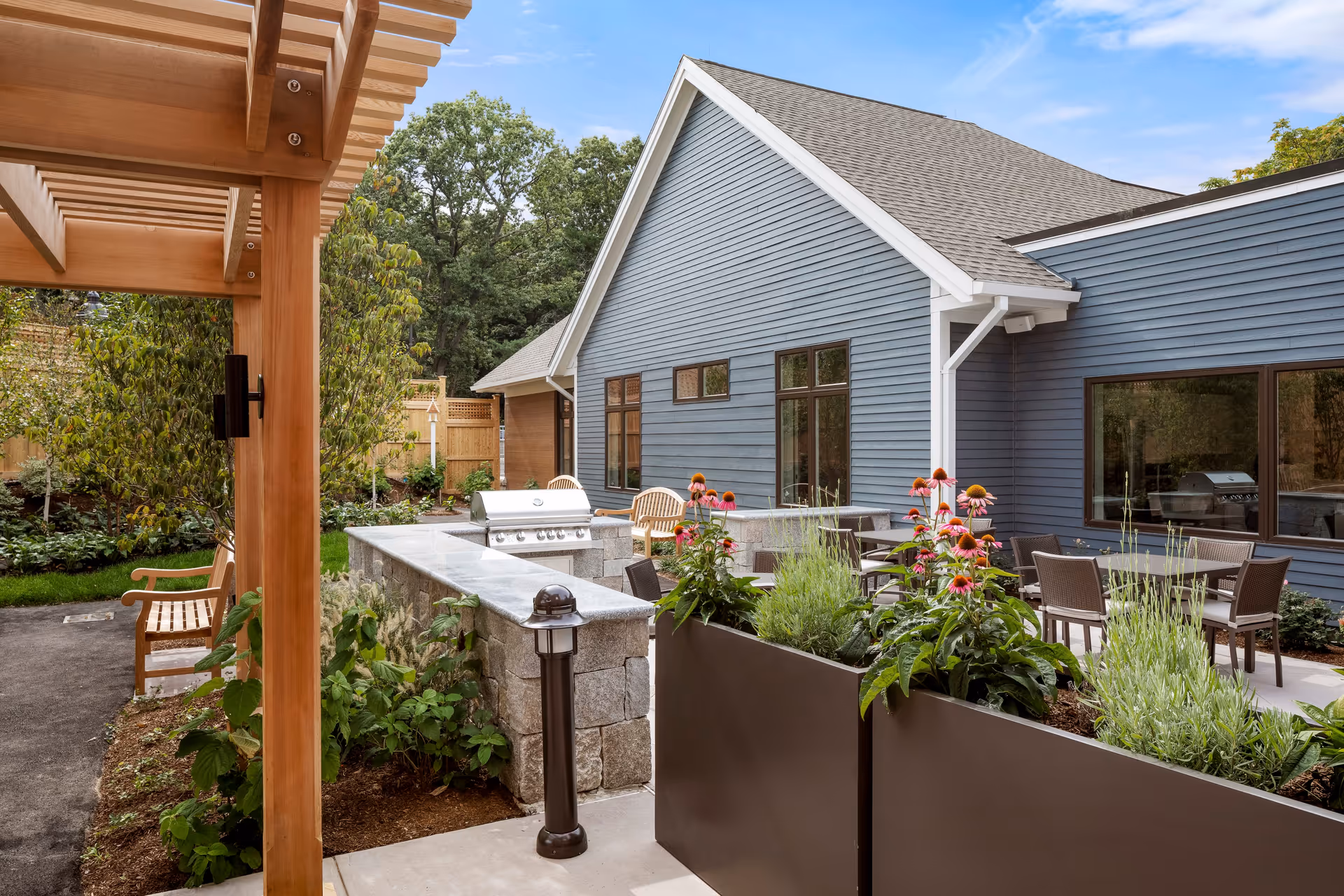 Outdoor patio area at a senior living facility featuring a stone countertop with a built-in grill, several tables and chairs for seating, raised planters with flowering plants, a wooden pergola, and a blue building with multiple windows in the background.
