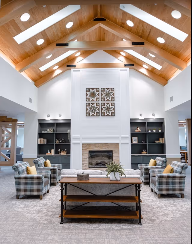 A spacious and well-lit living room area with a high vaulted wooden ceiling featuring skylights and circular windows. The room has a central fireplace with decorative wall art above it, flanked by built-in dark shelves filled with books and decor. There are four plaid armchairs with yellow cushions arranged around a wooden coffee table with a plant and decorative items on it. The carpet is light gray, and the overall decor is modern and cozy.
