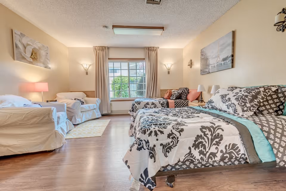 A bright and spacious bedroom with two beds featuring black and white patterned bedding. There are two white armchairs with a pink lamp on a side table between them on the left side of the room. A large window with beige curtains lets in natural light, and the walls are decorated with framed artwork. The floor is wooden, and the room has a cozy and inviting atmosphere.