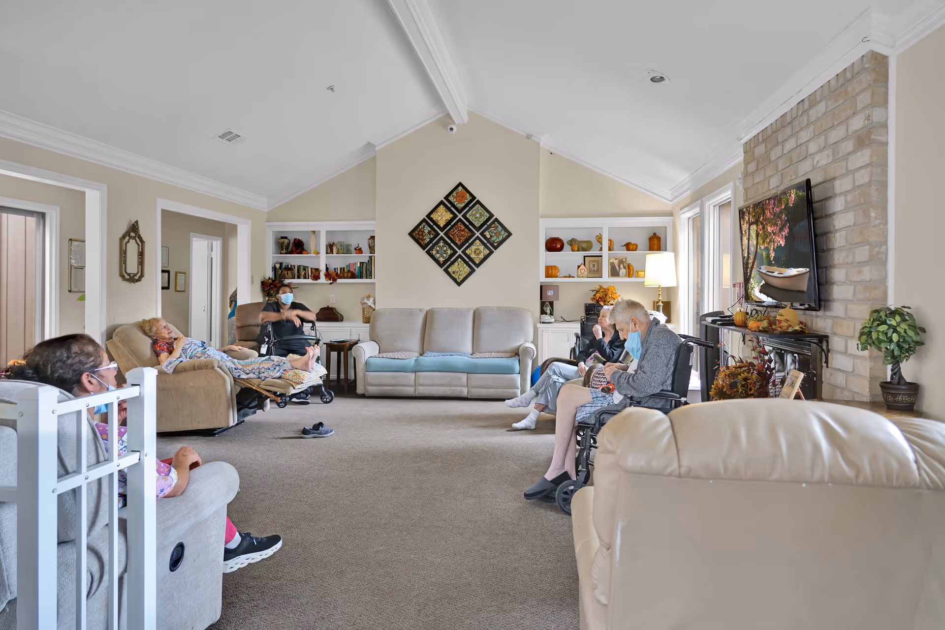 A spacious living room in a senior care facility with several elderly residents seated on recliners and wheelchairs, some wearing masks. The room features beige walls, a high vaulted ceiling, built-in shelves with decorative items, a wall-mounted TV above a brick fireplace, and a beige carpeted floor.