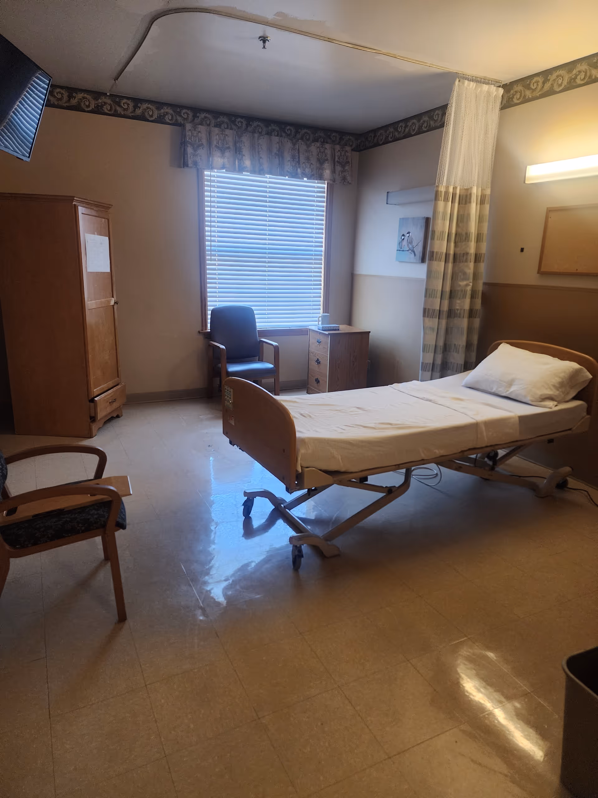 A senior care patient room with an adjustable hospital bed, chairs, a window with blinds, a bedside cabinet, and a privacy curtain.