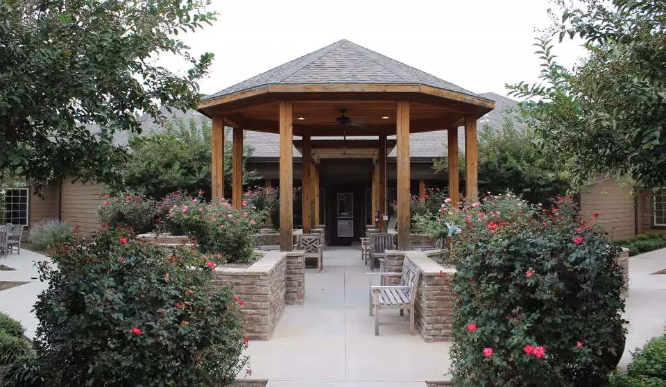 Outdoor courtyard area with a wooden gazebo structure in the center, surrounded by rose bushes and greenery. There are benches along the walkway leading to the gazebo, and the building exterior is visible in the background.
