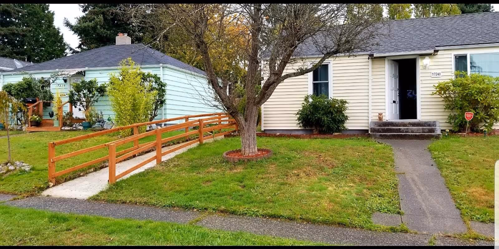Front exterior view of a single-story residential building with beige siding, a small porch with steps, a tree in the front yard, and a wooden ramp leading to the neighboring house with light blue siding. The lawn is green with some bushes near the houses.