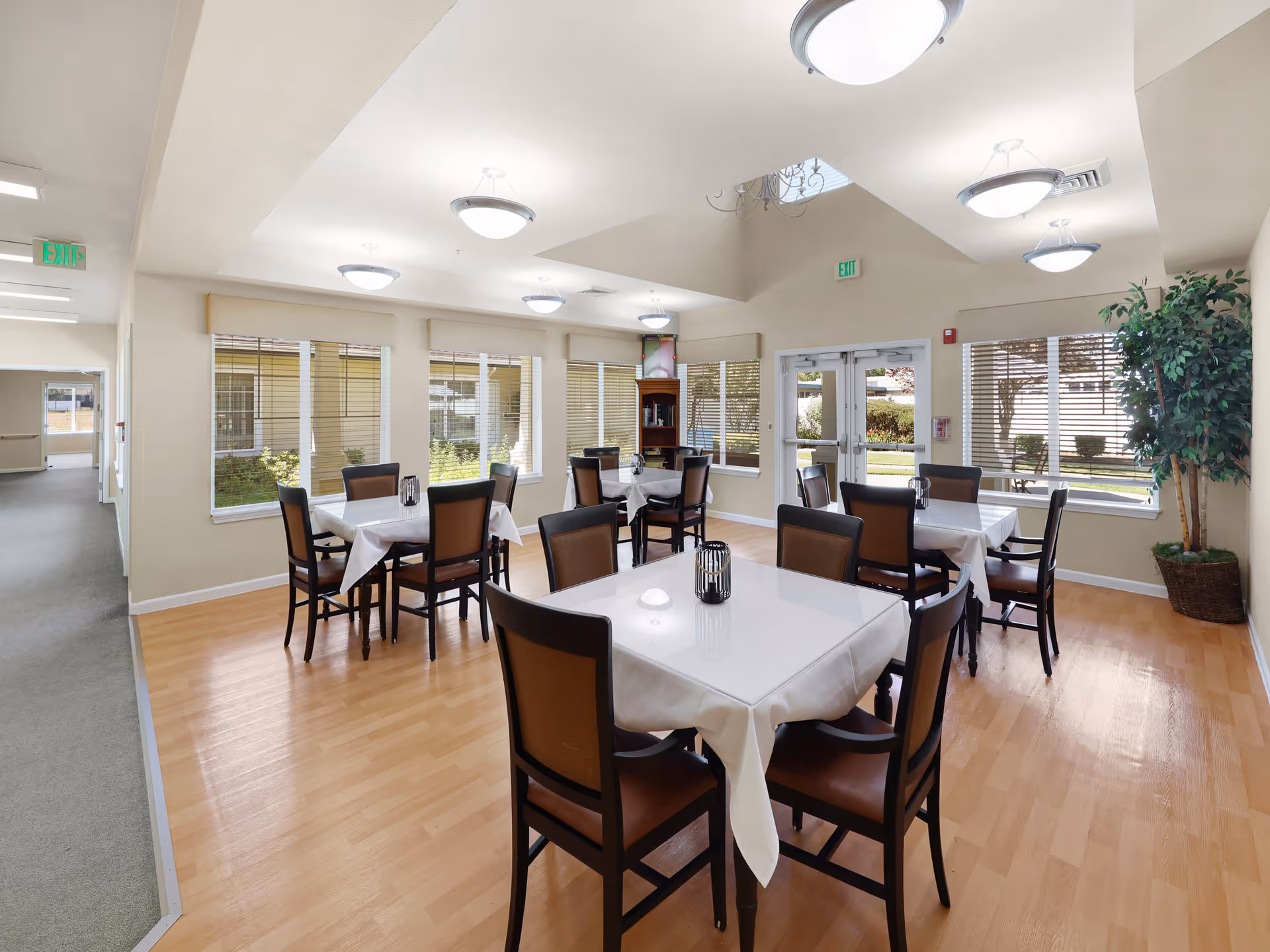 A bright dining room with multiple square tables covered with white tablecloths, each surrounded by four brown chairs. The room has large windows with blinds, light wood flooring, and ceiling lights. There is a potted plant in the corner and double glass doors leading outside.