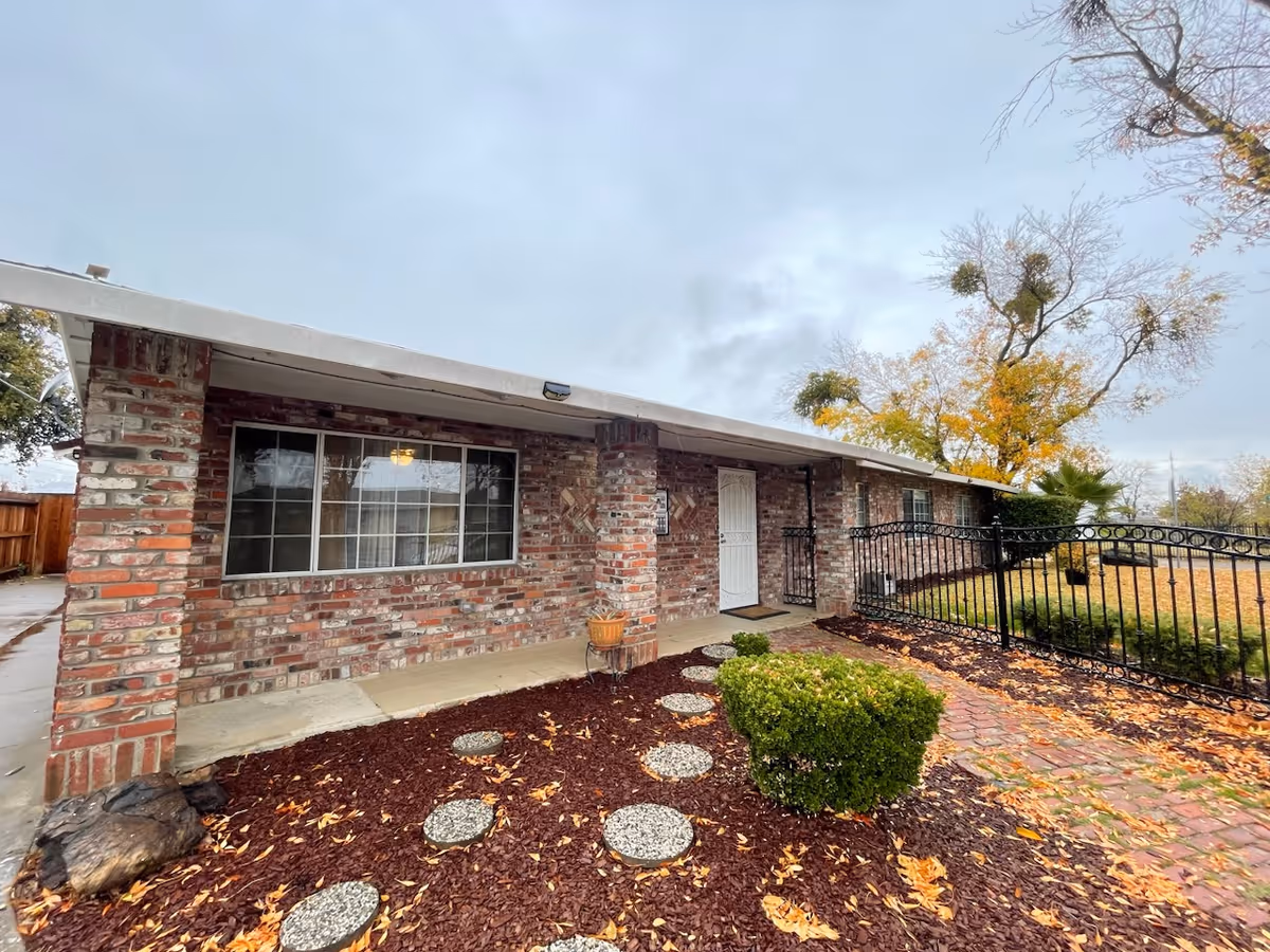 Front exterior of a single-story brick building with a covered porch, landscaped mulch bed with stepping stones, and an iron fence.