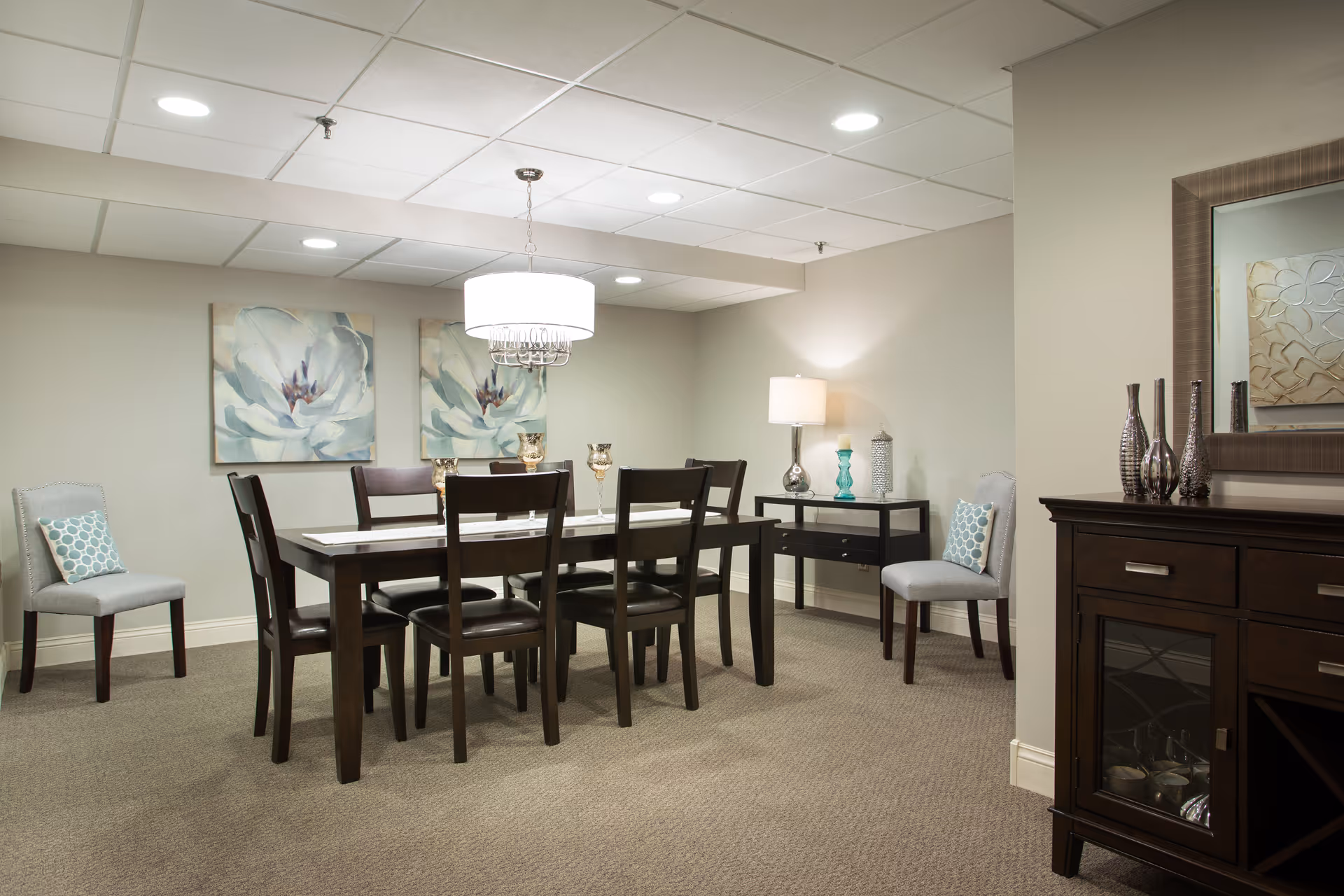A dining room with a dark wooden table surrounded by six matching chairs. Two additional upholstered chairs with patterned cushions are placed against the walls. The room features beige walls, carpeted floor, two large floral paintings, a modern chandelier above the table, a side table with a lamp and decorative items, and a dark wooden cabinet with a mirror above it.