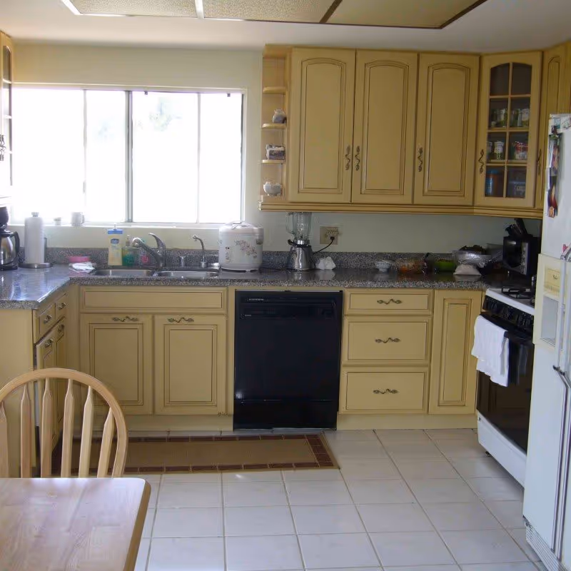 Sunlit kitchen with beige-yellow cabinets, granite countertops, a sink under a window, a black dishwasher, stove, and a wooden dining chair.