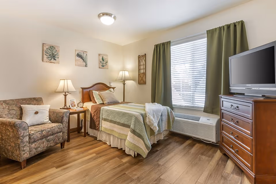 A cozy bedroom with a single bed covered in a green and beige quilt, a wooden headboard, and multiple pillows. Next to the bed is a wooden nightstand with a lamp, a clock, and a framed photo. There is a patterned armchair with a white pillow to the left of the nightstand. On the right side of the room, there is a wooden dresser with a flat-screen TV on top. A window with green curtains and white blinds is behind the bed, and the floor is wood.