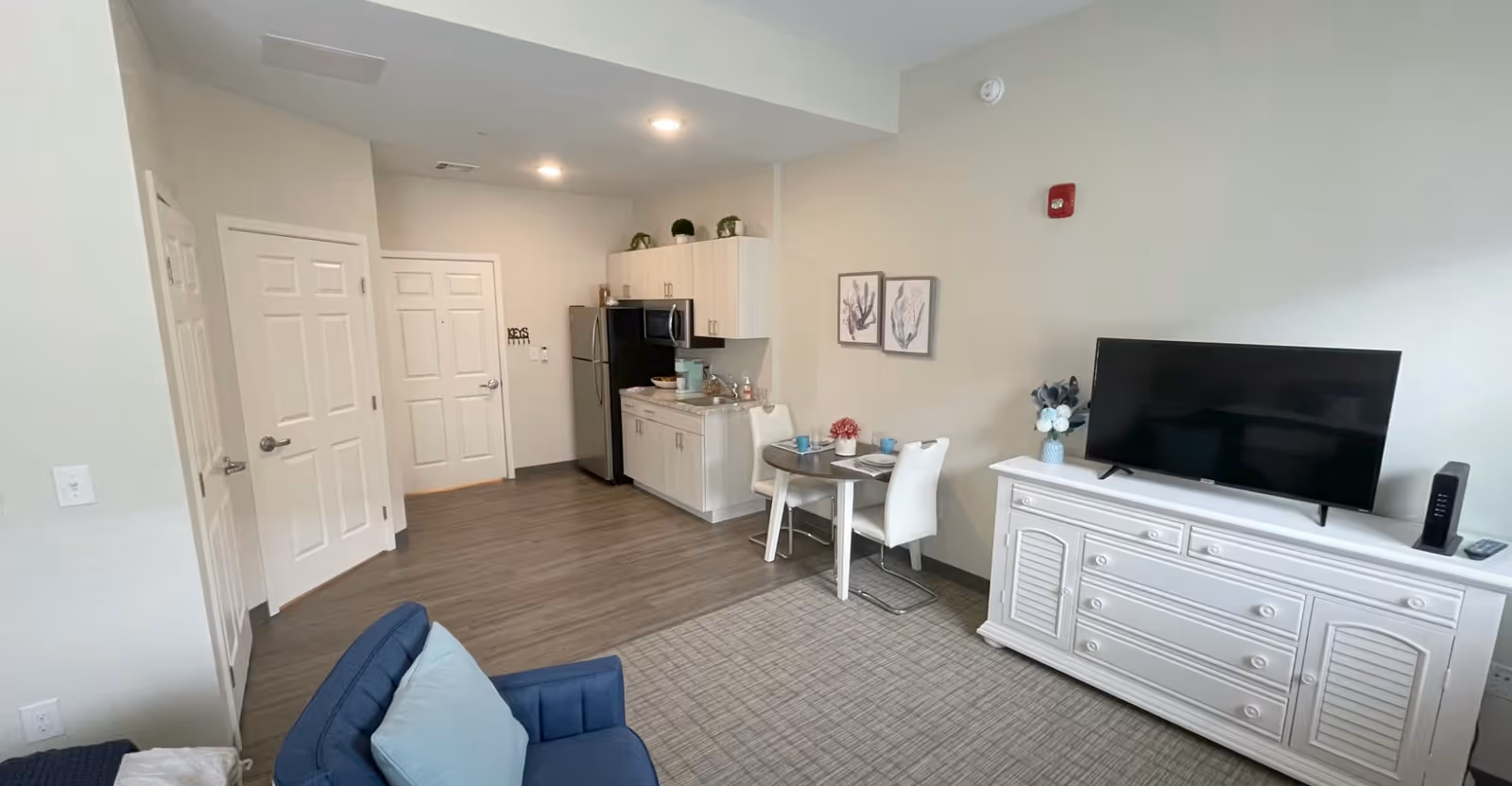 Interior view of a senior living community apartment featuring a small kitchenette with a refrigerator, microwave, and cabinets. A round dining table with two white chairs is set with blue cups and a small flower vase. A white dresser holds a flat-screen TV and a decorative vase with flowers. A blue armchair with a light blue pillow is partially visible in the foreground. The room has light-colored walls and wood flooring.