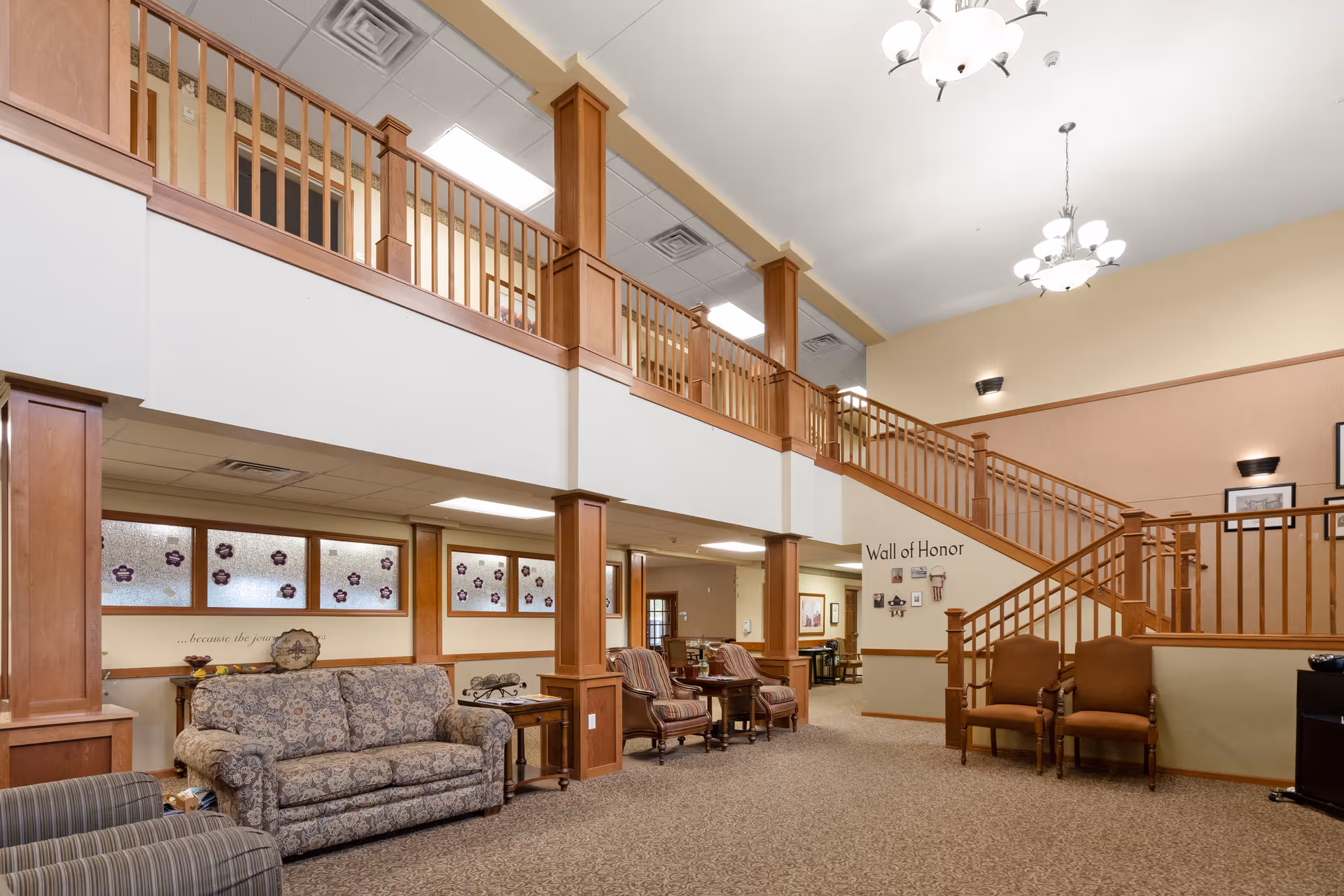 Spacious senior living facility common area with a high ceiling, wooden railings on the upper level, and a staircase. The room features comfortable seating including a patterned sofa, armchairs, and side tables. There is a 'Wall of Honor' display on the wall near the staircase. The area is well-lit with ceiling lights and wall sconces.