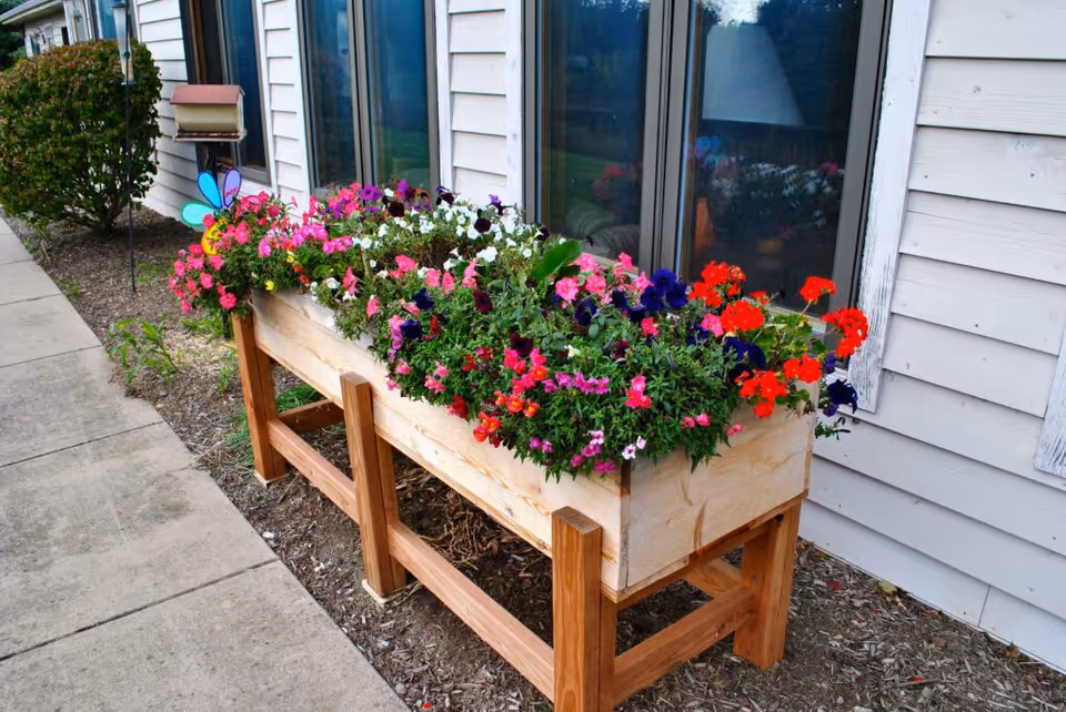 A raised wooden planter box filled with colorful flowers including pink, red, purple, and white blooms, placed outside next to a building with beige siding and windows. There is a concrete sidewalk on the left and some green bushes in the background.