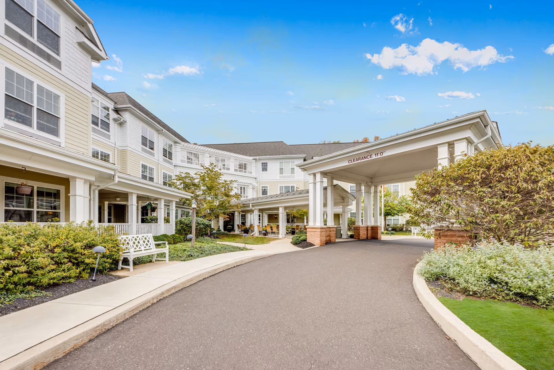 Exterior view of Sunrise of North Wales senior living facility showing a covered entrance with clearance sign, surrounded by well-maintained landscaping, bushes, and benches along the sidewalk under a blue sky with scattered clouds.