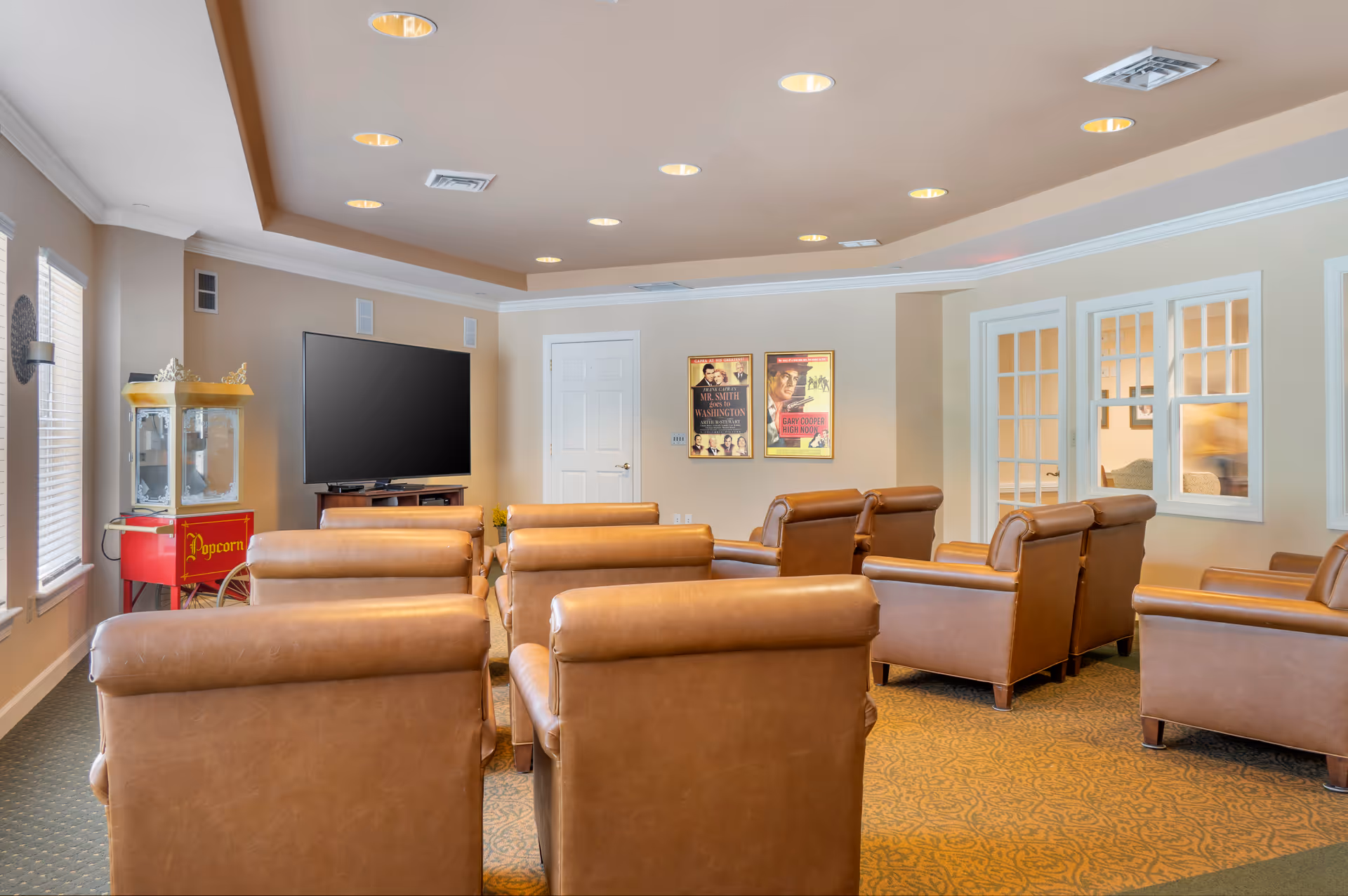 A cozy media room with several brown leather armchairs arranged in rows facing a large flat-screen TV mounted on a stand. To the left, there is a vintage-style popcorn machine. The room has beige walls, a patterned carpet, recessed ceiling lights, and two framed movie posters on the wall. Windows with white blinds and white trim are visible on the left and right sides.