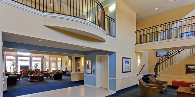 Two-story senior living facility lobby with seating areas, staircases, balcony railings, and an elevator.