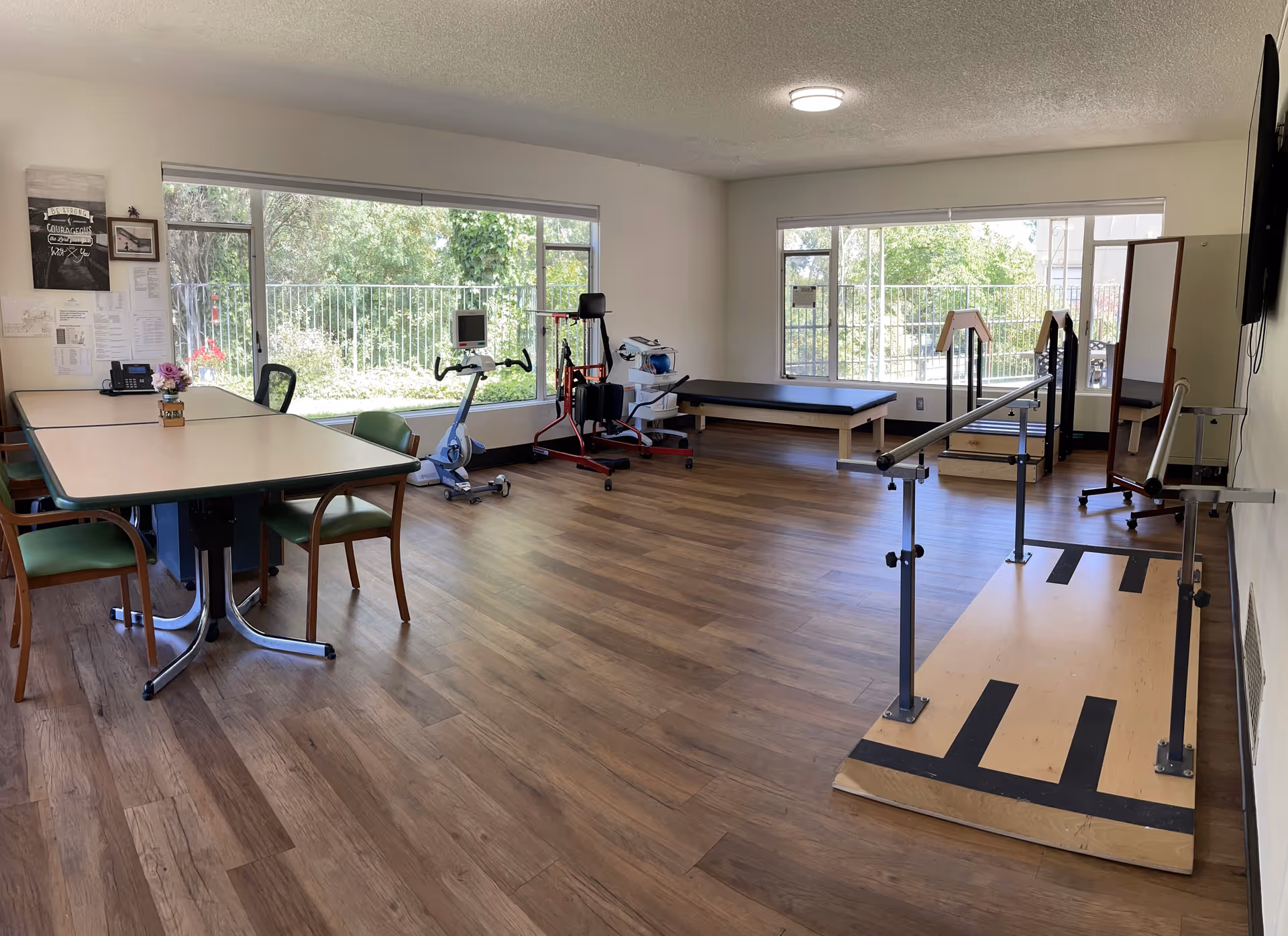 A spacious rehabilitation room with wooden flooring, large windows letting in natural light, exercise equipment including a stationary bike and parallel bars, a treatment table, and a table with chairs on the left side. The room has white walls and a TV mounted on the right wall.