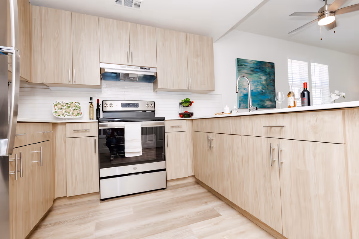 Modern kitchen with light wood cabinets, a stainless steel oven and refrigerator, a white countertop with a sink and faucet, a ceiling fan, and decorative items including bottles of wine, a wine glass, and a painting on the wall.