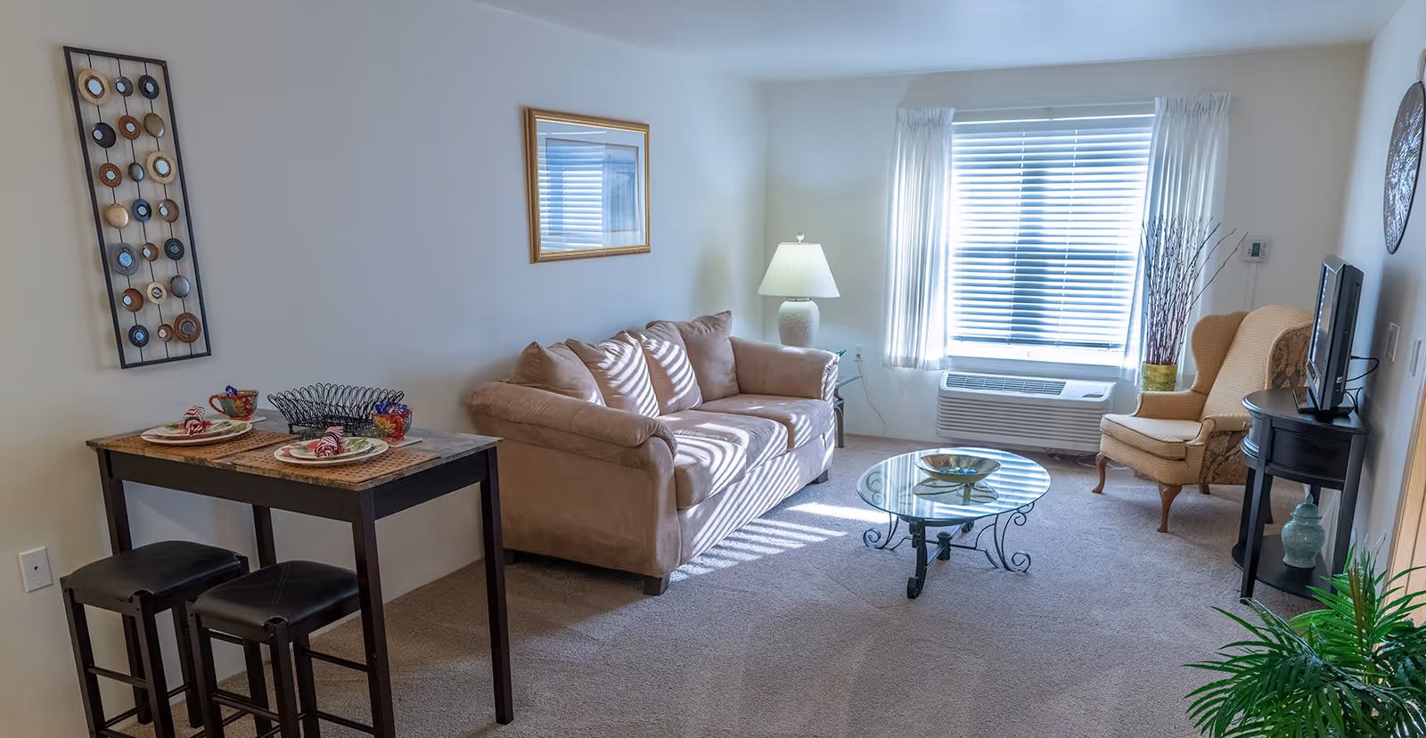A bright living room with a beige sofa, a glass coffee table, a beige armchair, a small dining table set for two with colorful cups and plates, a lamp on a side table, a window with white blinds and curtains, and decorative wall art.