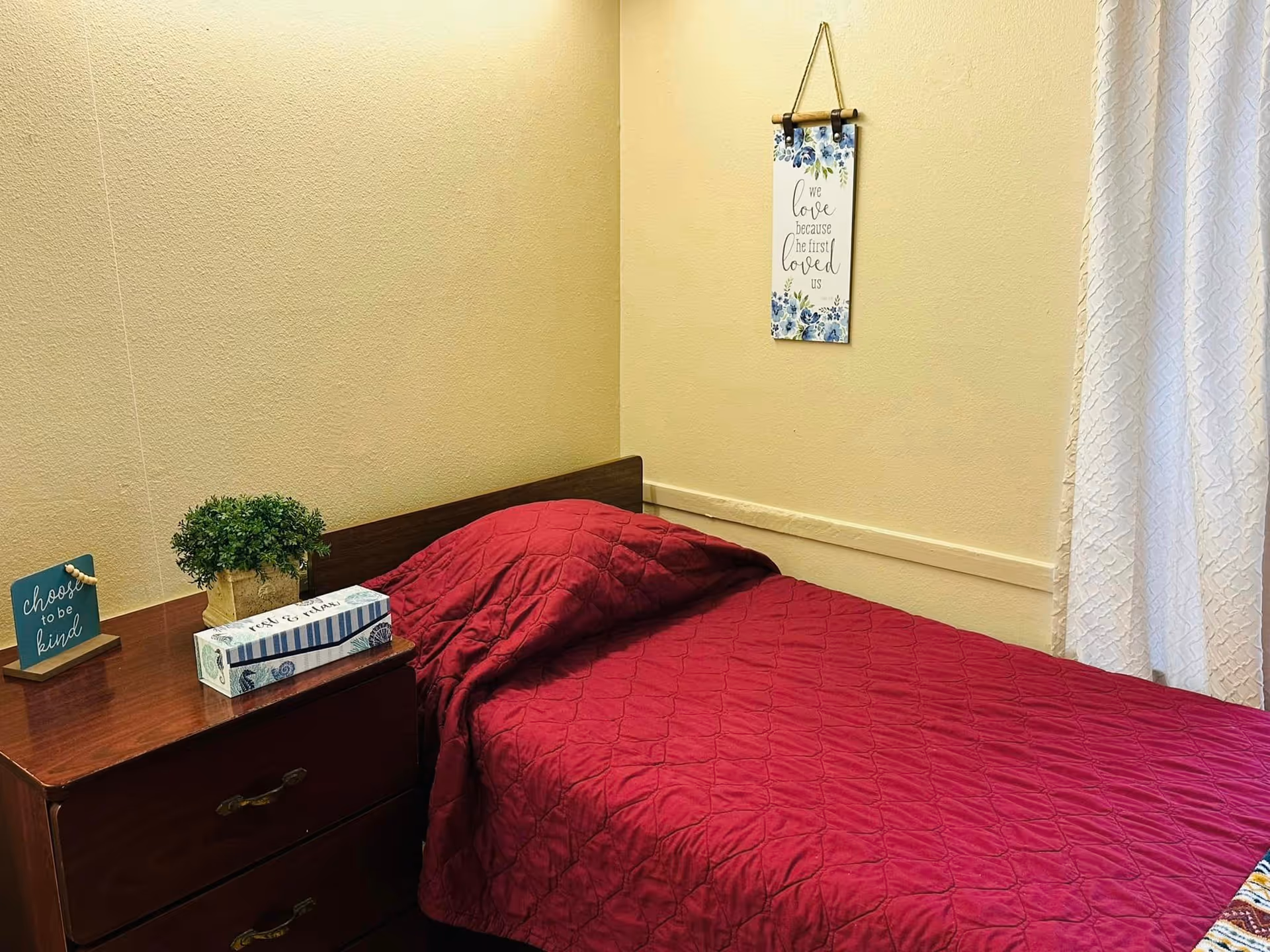 A small bedroom with a single bed covered in a red quilted blanket. Next to the bed is a wooden nightstand with a small potted plant, a decorative box of tissues, and a small sign that reads 'choose to be kind'. On the wall above the bed hangs a decorative sign with blue flowers and the text 'we love because he first loved us'. White curtains are partially visible on the right side.