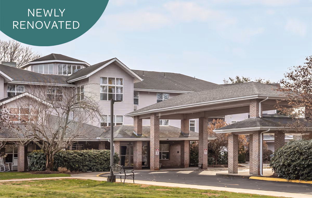 Exterior view of a multi-story senior living facility with a covered entrance supported by brick pillars. The building has multiple windows and a sloped roof. There is a bench and some landscaping with bushes and trees in front of the building. A green circular graphic in the top left corner says 'NEWLY RENOVATED'.