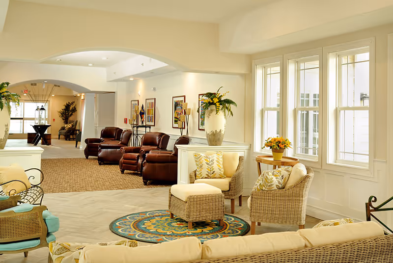 A bright and spacious senior living facility common area with large windows letting in natural light. The room features comfortable wicker and cushioned chairs with patterned pillows, a round colorful rug, and a small wooden table with a flower pot. In the background, there are leather armchairs, framed artwork on the walls, and decorative plants, creating a welcoming and cozy atmosphere.