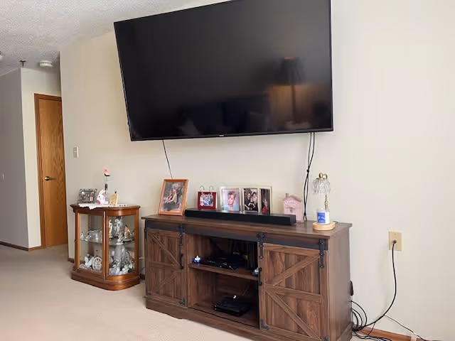 A living room area featuring a large flat-screen TV mounted on a beige wall above a wooden entertainment cabinet with barn-style doors. The cabinet holds various electronic devices and is decorated with framed photos and a small lamp. To the left, there is a small wooden display cabinet with glass doors containing decorative items. A closed wooden door is visible in the background.