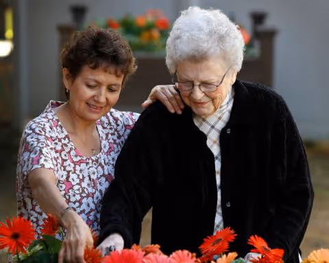An elderly woman with white hair and glasses is being assisted by a younger woman with short brown hair as they tend to a garden with red and orange flowers outdoors.