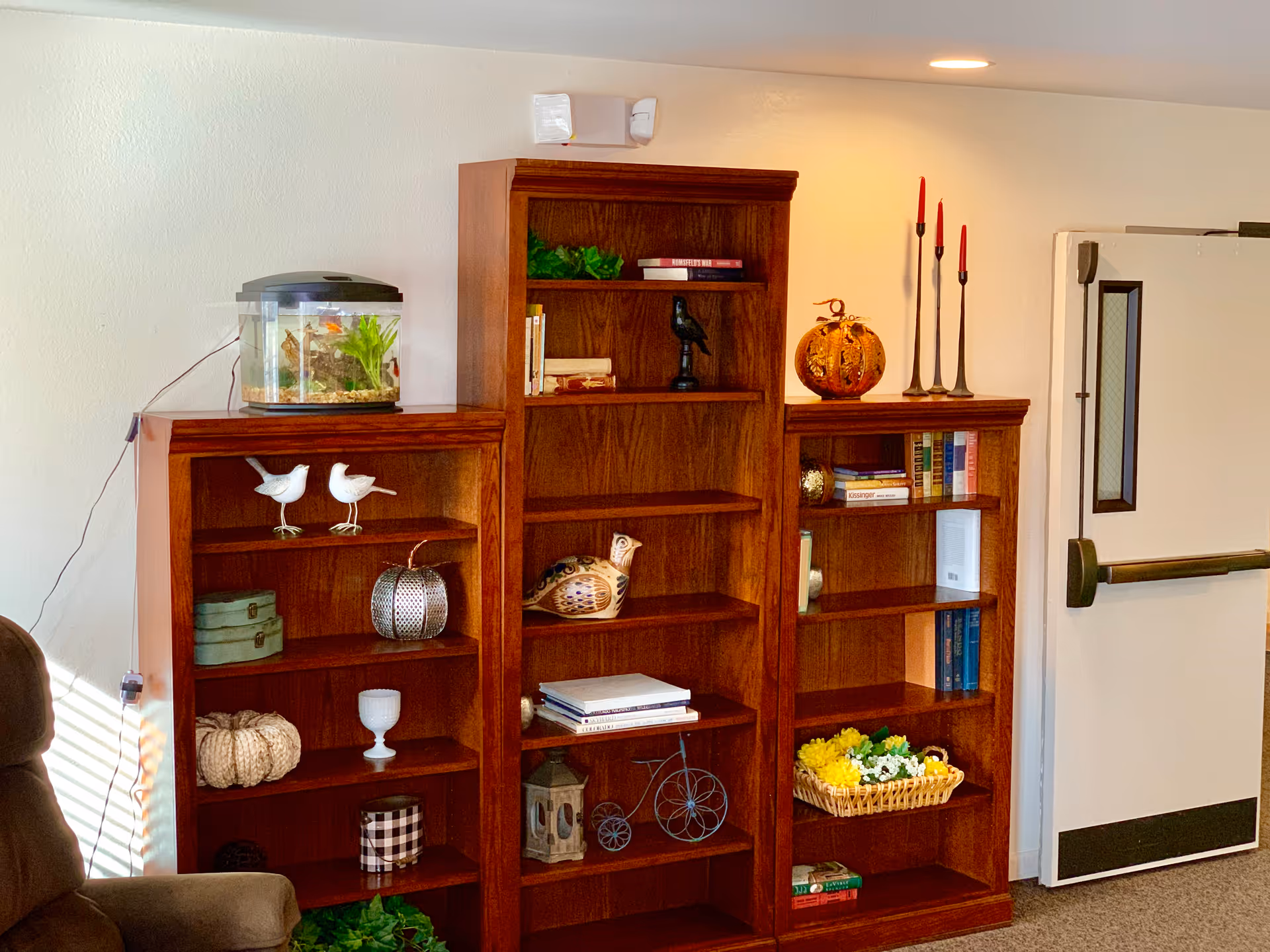 Three wooden bookshelves arranged side by side against a white wall. The shelves hold various decorative items including books, small sculptures, a fish tank, candles, and a basket of flowers. To the right is a white door with a horizontal push bar. Part of a brown armchair is visible on the left side of the image.