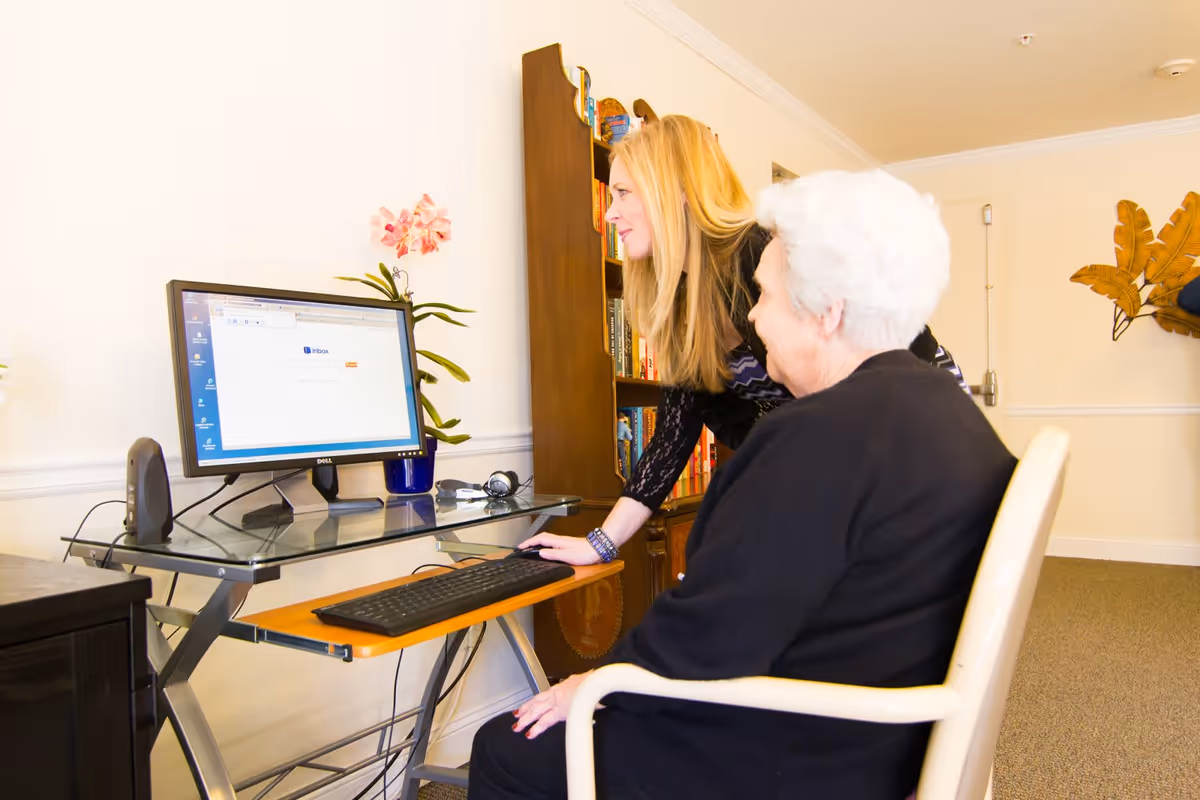 An elderly woman with white hair sitting in a chair at a glass computer desk, while a younger woman with long blonde hair leans over to assist her with the computer. The room has beige walls, a bookshelf filled with books, and decorative plants on the wall.