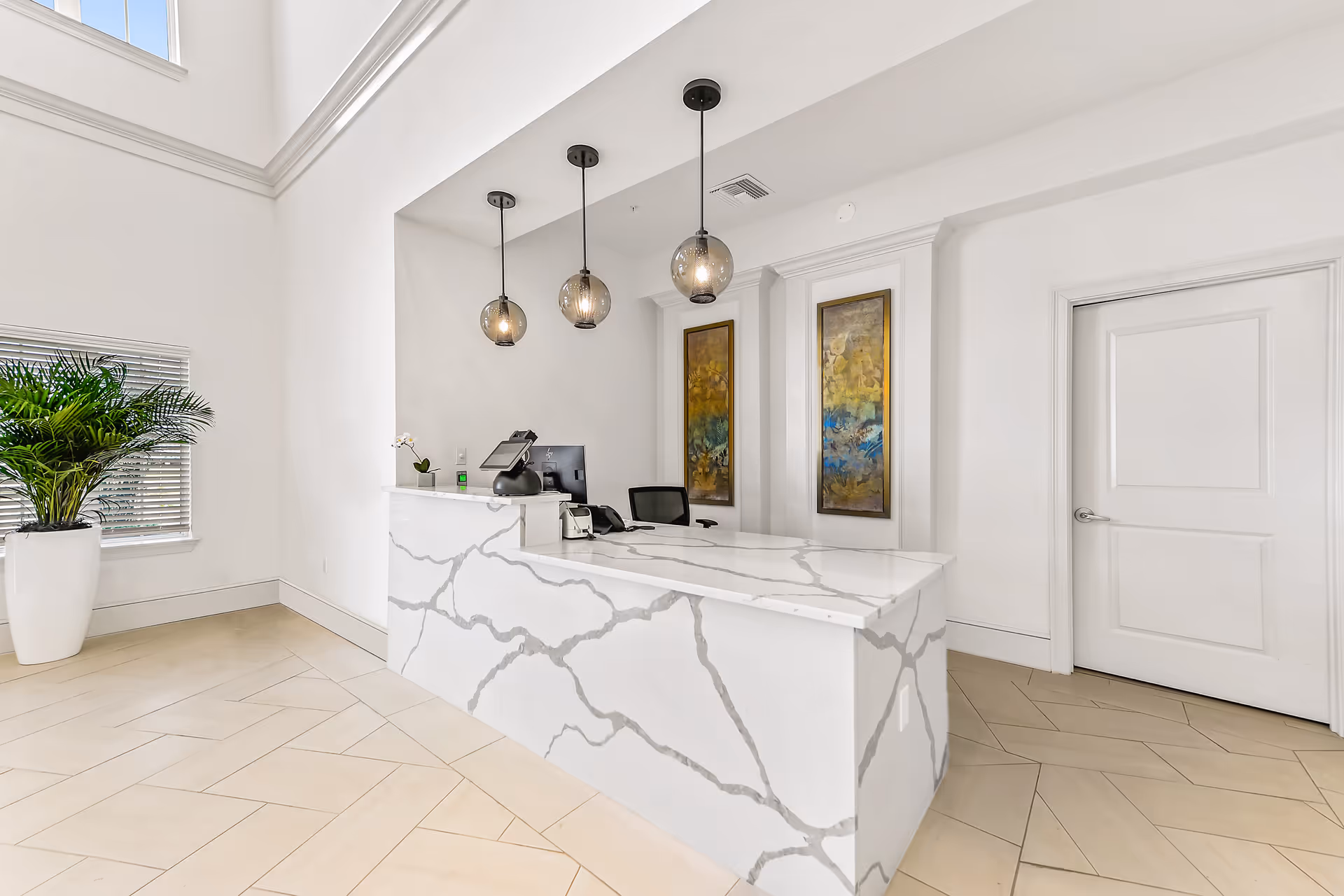 A modern reception desk area with a white marble counter featuring gray veining. Behind the desk are two framed abstract paintings with gold, blue, and green tones. Three pendant lights with round glass shades hang above the desk. To the left, there is a large potted plant near a window with blinds. The floor is tiled in a light beige color with a herringbone pattern, and a white door is visible on the right side of the image.