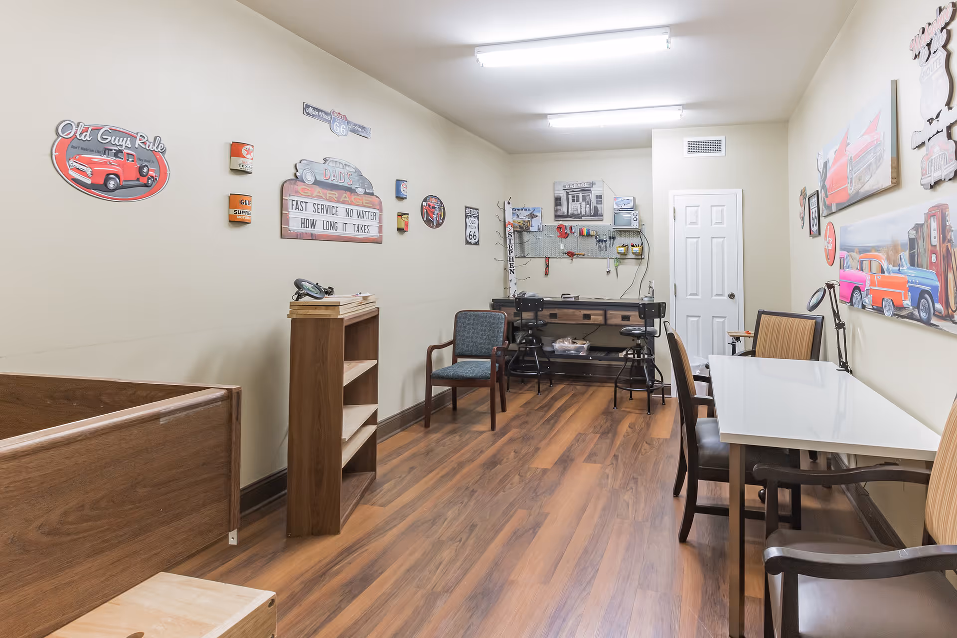 Long interior activity room with tables, chairs, a workbench and vintage car wall decorations.