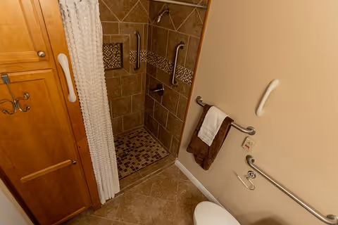A senior-friendly bathroom featuring a walk-in tiled shower with grab bars, a white textured shower curtain, a wooden cabinet with hooks, a towel rack holding brown and white towels, and additional grab bars near the toilet area.