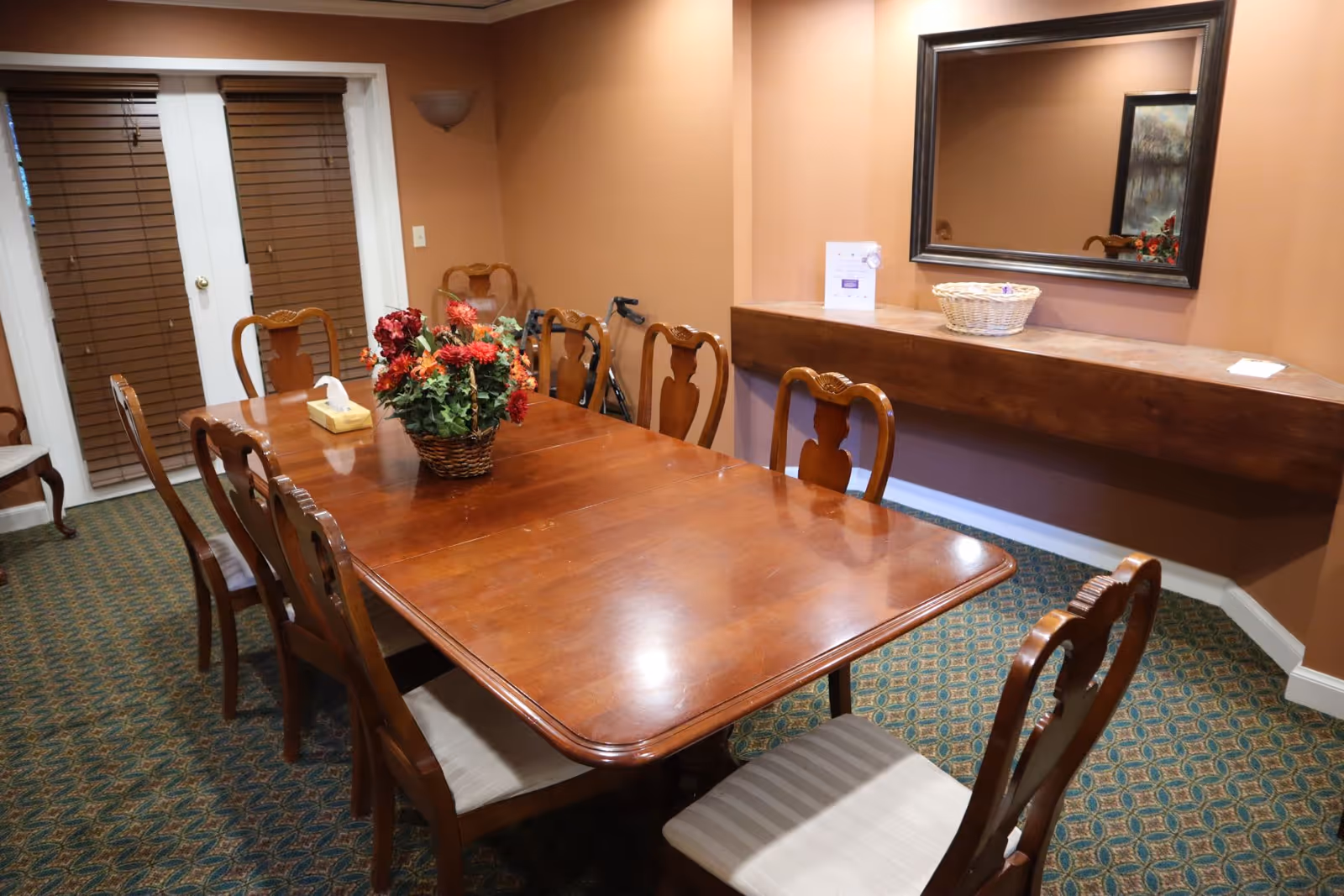 A dining room with a long wooden table surrounded by eight wooden chairs with cushioned seats. A basket of red flowers is placed in the center of the table. The room has patterned carpet flooring, beige walls, a large mirror on one wall above a wooden ledge, and French doors with closed blinds.