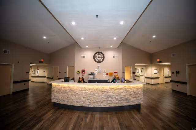 Curved stone-front reception desk in a spacious lobby with staff at the desk, a large wall clock on the back wall, and hallways leading off to either side.