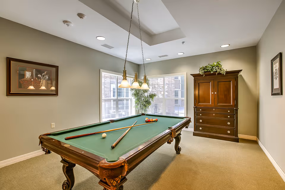 A well-lit room with a green felt pool table in the center, two pool cues resting on the table, and a set of billiard balls arranged for a game. The room has beige walls, a large window letting in natural light, a wooden cabinet with drawers and a plant on top, and framed artwork on the walls. A hanging light fixture with three lamps is positioned above the pool table.