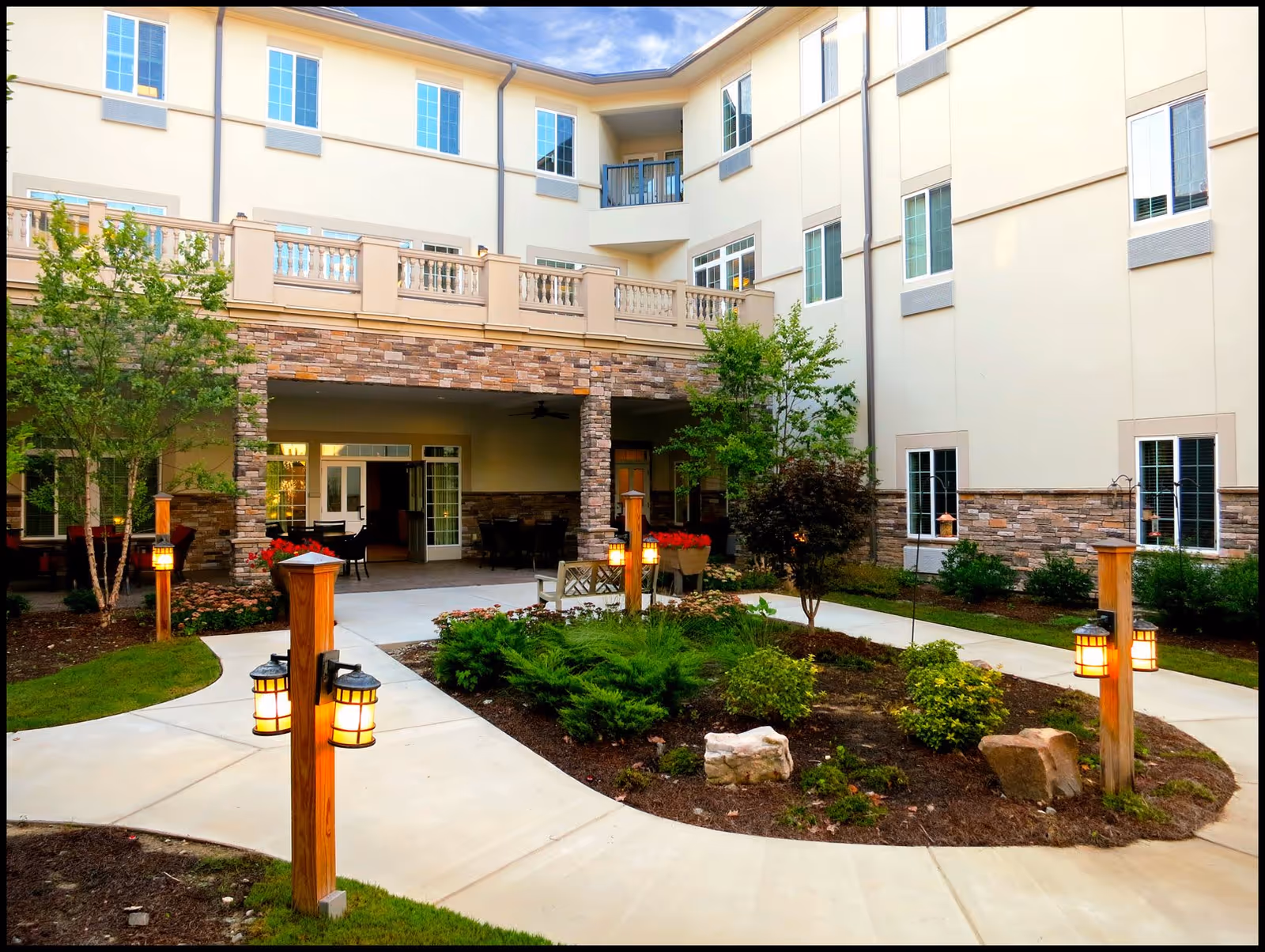Outdoor courtyard area of The Cambridge - Assisted Living & Memory Care facility featuring a landscaped garden with shrubs, small trees, rocks, and multiple wooden posts with lantern-style lights. The courtyard is surrounded by a multi-story building with beige walls, stone accents, and numerous windows. There is a covered patio area with tables and chairs visible in the background.