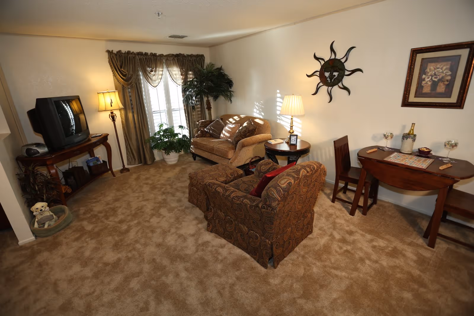 A cozy living room with beige carpet, a patterned armchair, a loveseat, a wooden side table with a lamp, a small dining table with two chairs, and a TV on a wooden stand. There are curtains on the window, a potted plant, wall decorations including a sun sculpture and framed artwork, and a small dog bed with a stuffed animal in the corner.