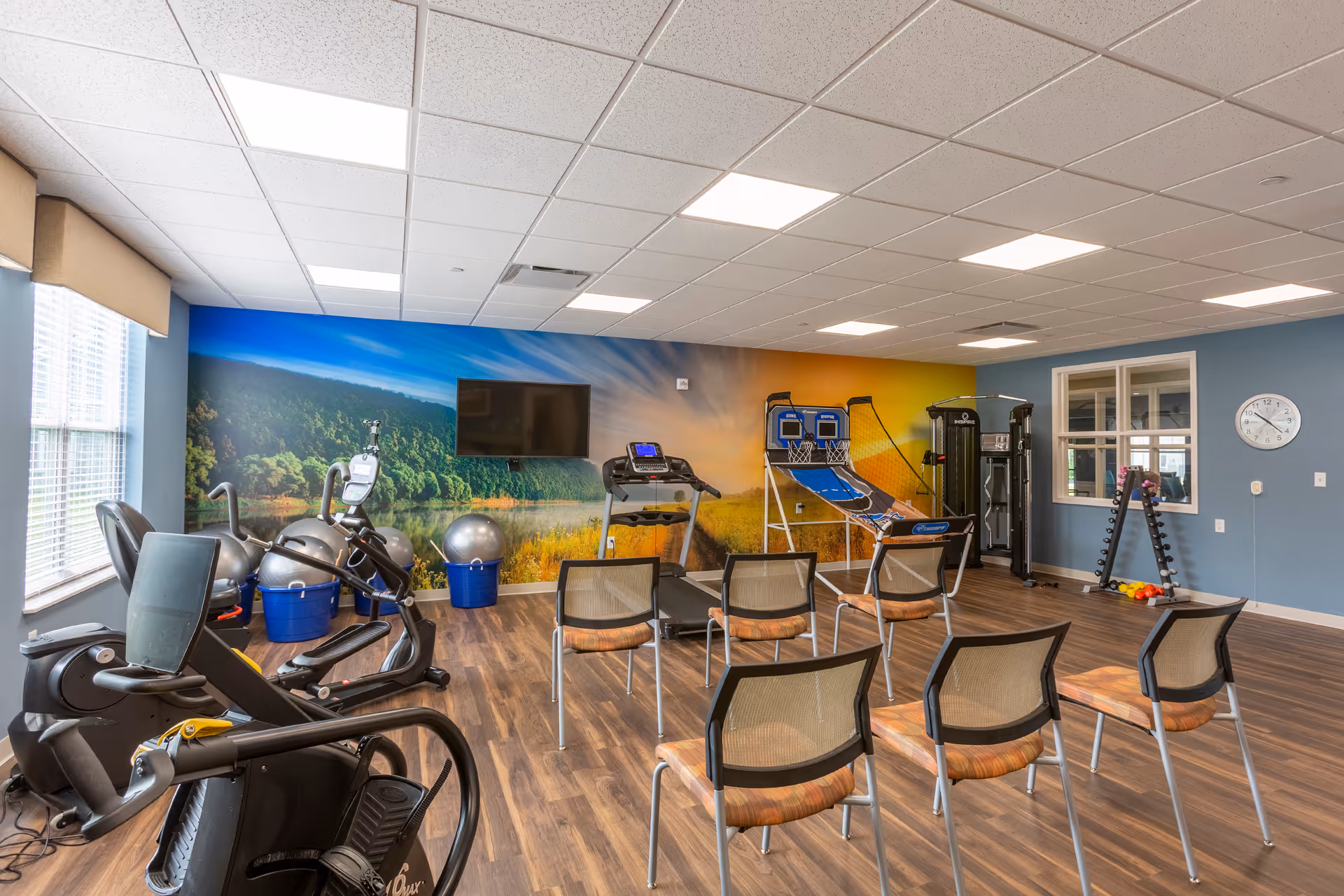 A fitness room with exercise equipment including stationary bikes, a treadmill, exercise balls, a basketball shooting game, and a weight machine. There are six chairs arranged in two rows facing a wall-mounted TV. The room has wood flooring, blue walls, and a large window letting in natural light. A colorful mural of a nature scene decorates one wall.