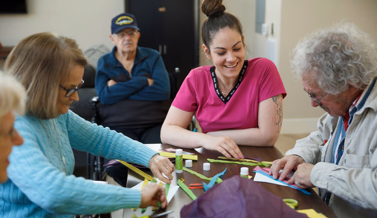 A group of elderly people and a caregiver sitting around a table engaged in a craft activity with colorful paper strips and glue sticks. The caregiver is smiling and interacting with the participants.