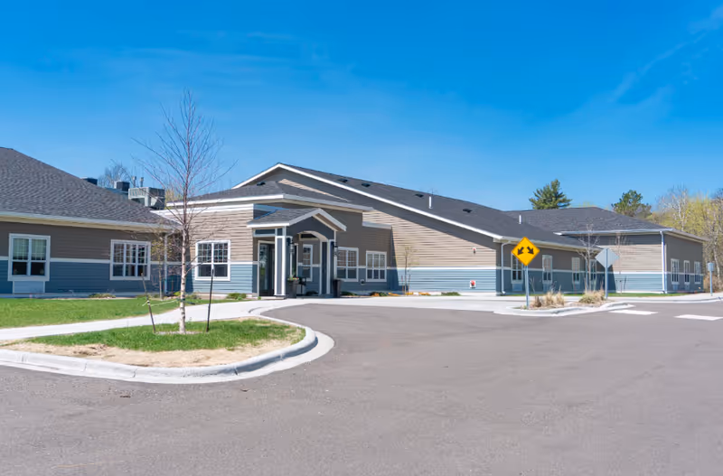 Single-story senior living facility building with a covered entrance, surrounding lawn, and a paved driveway under a clear blue sky.