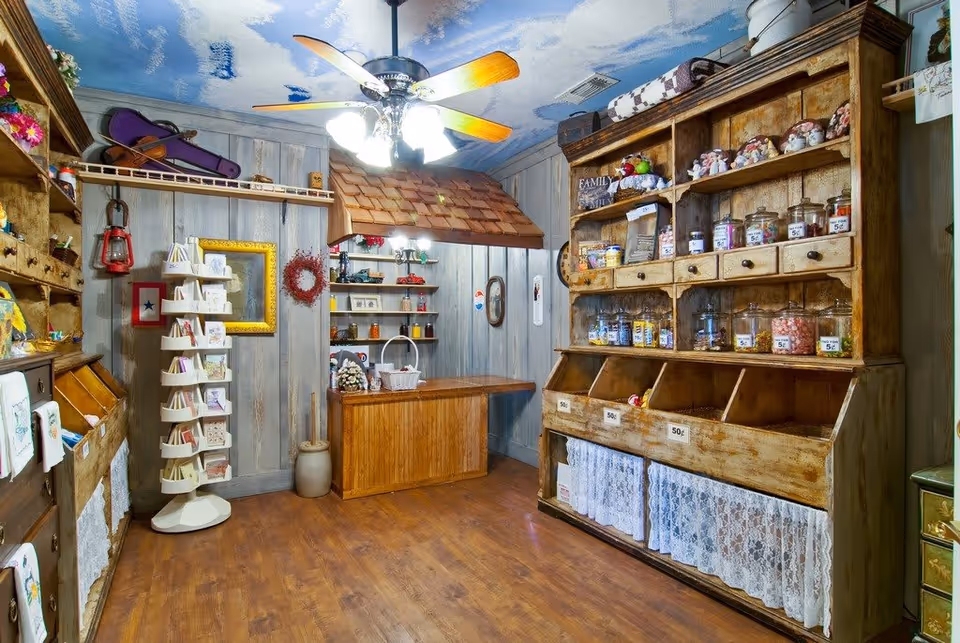 Interior of a rustic-style room with wooden shelves and cabinets displaying various jars, small items, and cards. The ceiling is painted with a sky and clouds motif, and a ceiling fan with wooden blades and lights hangs in the center. The room has wooden flooring and light-colored wooden walls, creating a cozy, vintage shop-like atmosphere.