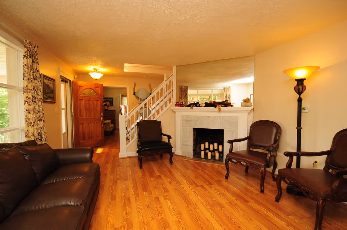 A cozy living room with wooden flooring, a dark brown leather sofa on the left, three dark wooden armchairs with leather cushions arranged near a white fireplace filled with candles. A tall floor lamp with a yellow shade is on the right side. The room has a large mirror above the fireplace, floral curtains on the window, and a wooden front door that is open, revealing another room and a staircase with a stairlift.
