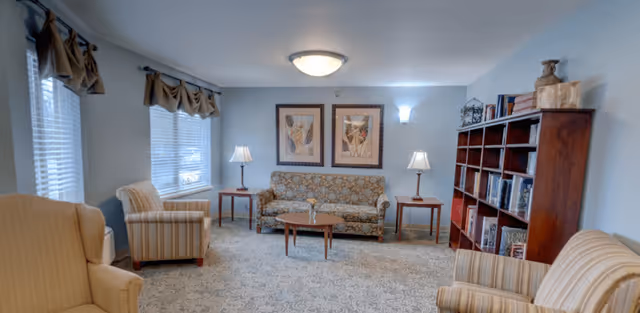 A cozy living room with a floral patterned sofa centered against the back wall, flanked by two wooden side tables each holding a lamp. Two framed artworks hang above the sofa. There are two striped armchairs and one solid beige armchair arranged around a small wooden coffee table. A large wooden bookshelf filled with books is on the right side of the room. The room has light blue walls, carpeted floor, and two windows with valance curtains on the left side.