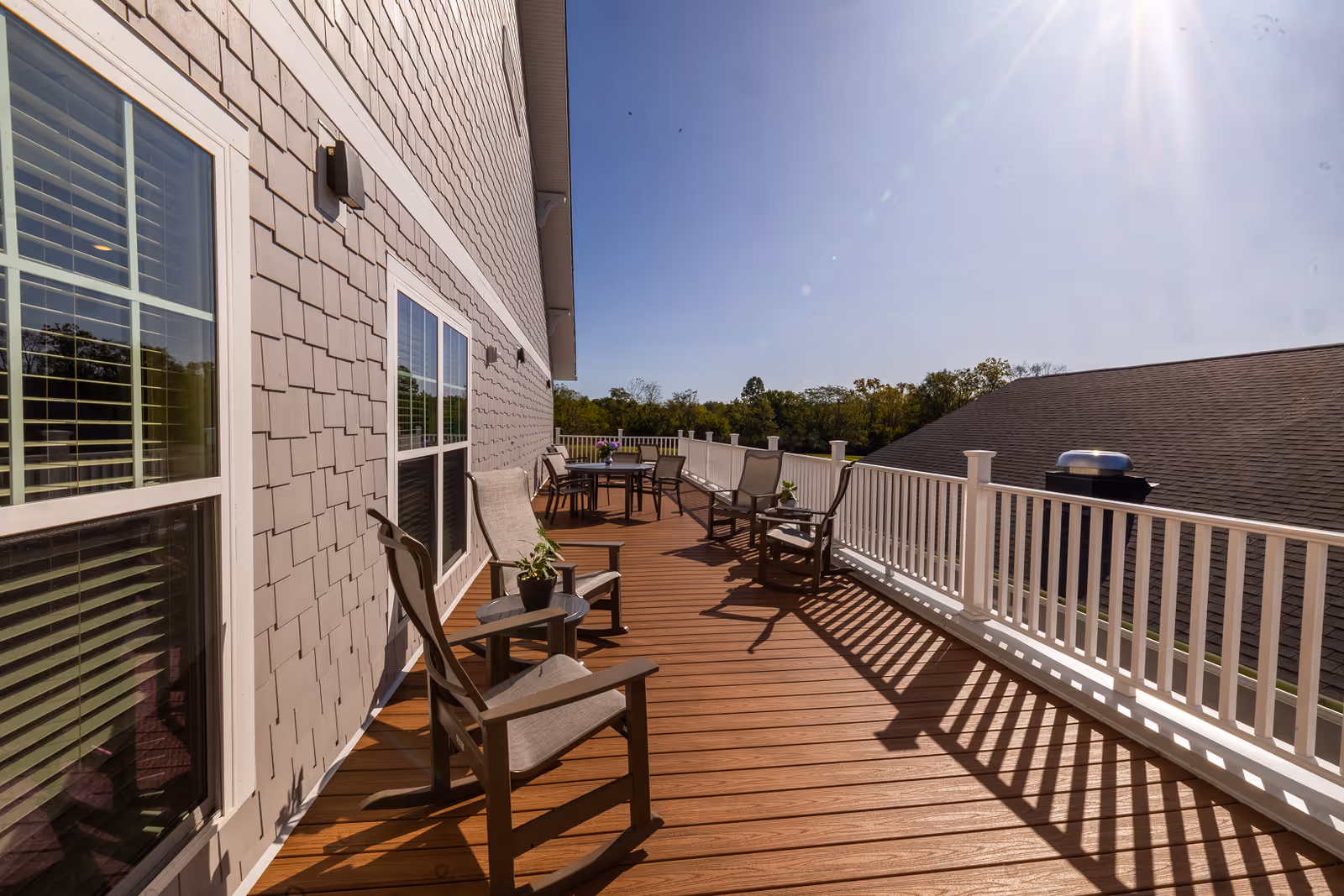 A spacious outdoor wooden deck attached to a building with beige siding and white-framed windows. The deck features several chairs, small tables with potted plants, and a round table with chairs in the background. A white railing lines the edge of the deck, and trees are visible in the distance under a clear blue sky with bright sunlight.