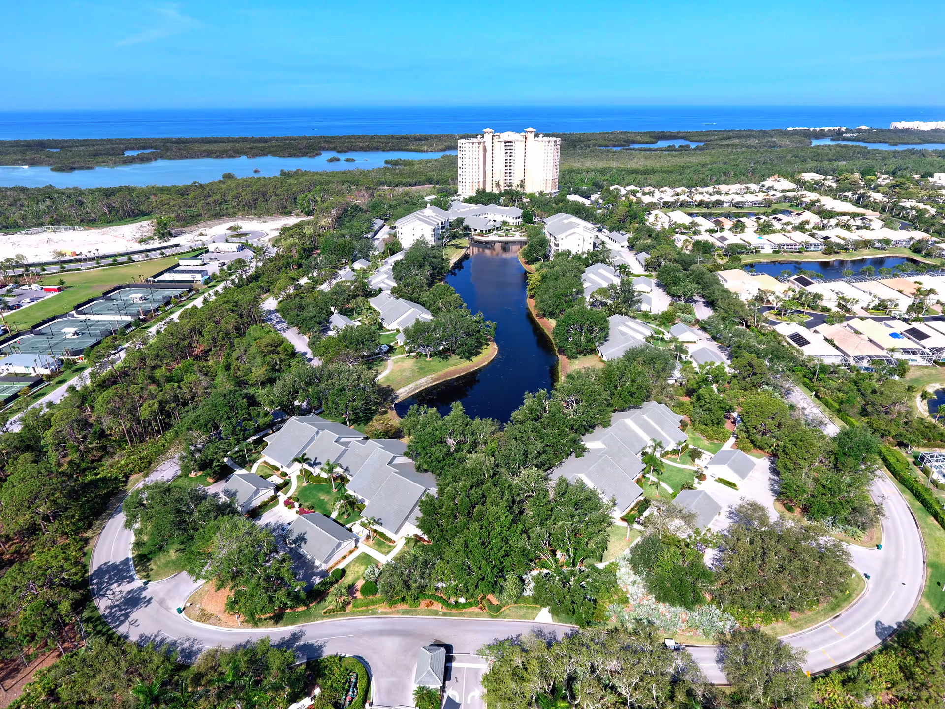 Aerial view of Arbor Trace senior living facility showing multiple residential buildings arranged around a central pond with lush greenery and trees. Roads curve around the property, and there are tennis courts visible on the left side. In the background, there are more residential areas, water bodies, and a clear blue sky.